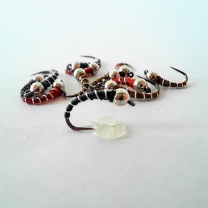 May include: A close-up of several fly fishing lures with silver beads and colorful wire wraps. The lures are arranged on a white surface.