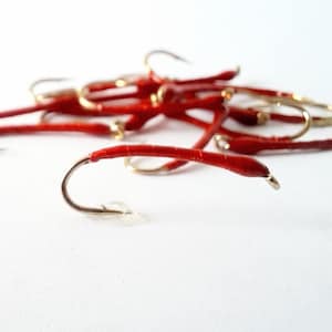 May include: A collection of red fishing flies with gold hooks. The flies are a vibrant red color and have a curved hook design. The image is taken against a white background, highlighting the details of the flies.