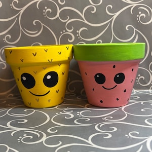 May include: Two small painted terracotta flower pots. One pot is yellow with a smiling face and a cactus pattern. The other pot is pink with a smiling face and a watermelon pattern.