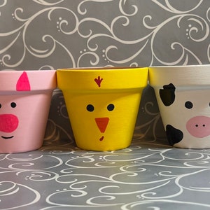 May include: Three terracotta flower pots painted with animal faces. The pots are painted pink, yellow, and white with black spots. The faces are a pig, a chick, and a cow.