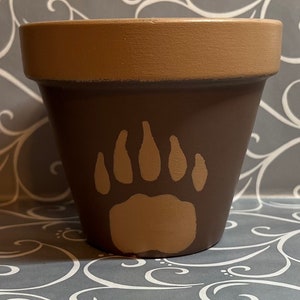 May include: A brown terracotta planter with a painted brown bear paw print in a lighter brown color.