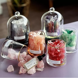 May include: Several glass containers filled with colourful raw crystals, some covered by glass domes. Each container features a VANT KEVIN label. A small glass dropper bottle labelled VANT KEVIN CRYSTAL STONE sits among loose pink crystals.