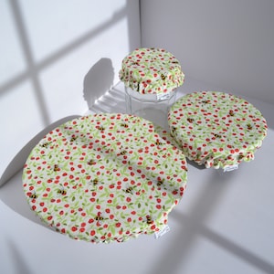 May include: Three reusable food covers with a white background and a pattern of red berries and yellow and black bees. The covers are made of fabric and have a ruffled edge.