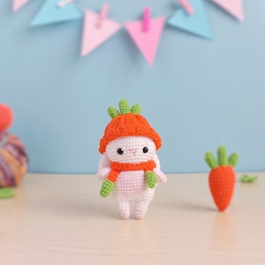 以下が含まれることがあります： オレンジ色の帽子とマフラーをつけた白い編みぐるみウサギ。ウサギは明るい木製の表面に立っており、右側に緑色の編みぐるみニンジンがある。