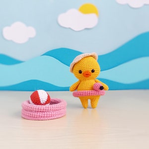 Może przedstawiać: Żółte, dziergane kurczątko w różowej dmuchanej oponce i białym, dzierganym kapeluszu. Kurczątko stoi na drewnianej powierzchni przed niebiesko-białym papierowym tłem z chmurami i słońcem.