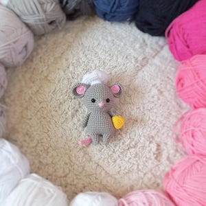 Może przedstawiać: Szara mysz wykonana na szydełku w białym czepku szefa kuchni i trzymająca żółty przedmiot wykonany na szydełku. Myszkę otaczają kłębki włóczki w różnych kolorach, w tym białym, różowym i szarym.
