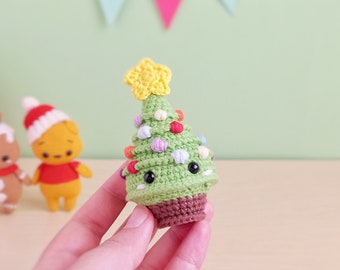 May include: A green crocheted Christmas tree with a yellow star on top and colorful ornaments. The tree has a brown crocheted base and a cute face with black eyes.
