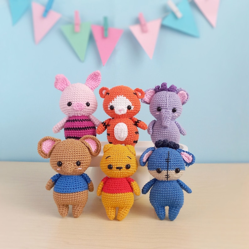 May include: A collection of six crocheted stuffed animals, including a pink pig, an orange tiger, a purple elephant, a brown elephant, a yellow bear, and a blue bear. The animals are all sitting on a light brown surface.