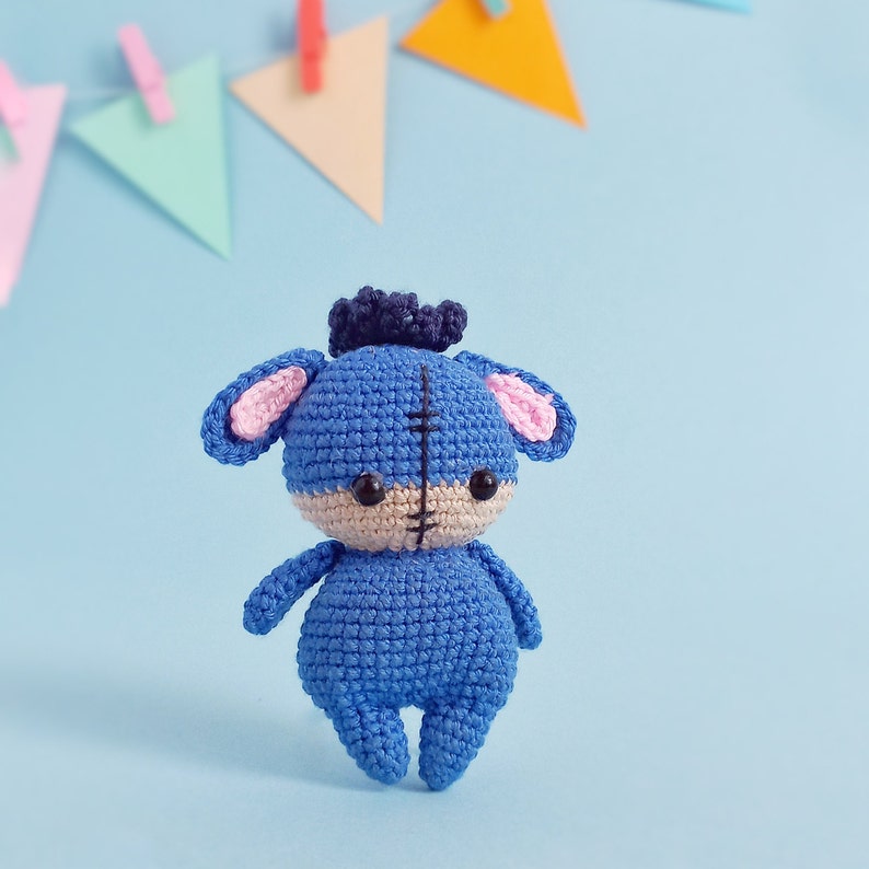 May include: A blue crocheted stuffed animal with pink ears and a black stitched face. The toy is standing on a light blue background.