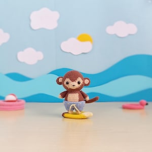 Puede incluir: Un mono marrón tejido a ganchillo con pantalones cortos azules, de pie sobre una tabla de surf amarilla. El mono tiene una cara y orejas de color canela. El fondo presenta una escena oceánica azul con nubes blancas y un sol amarillo.