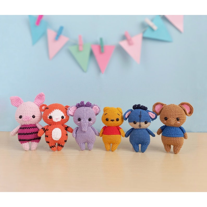 May include: Six crocheted stuffed animal toys, including Piglet, Tigger, Eeyore, Winnie the Pooh,  a blue donkey, and a brown bear. The toys are arranged in a row on a wooden surface.
