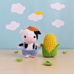 Puede incluir: Una figura de vaca tejida a ganchillo con manchas blancas y negras, con un peto azul y un sombrero pequeño. Junto a ella, una mazorca de maíz tejida a ganchillo con hojas verdes. El fondo presenta nubes de papel y un sol amarillo.