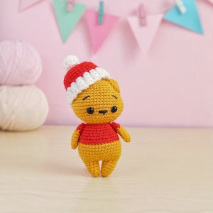 May include: A small crocheted teddy bear wearing a red and white Santa hat. The bear is standing on a light wood surface.