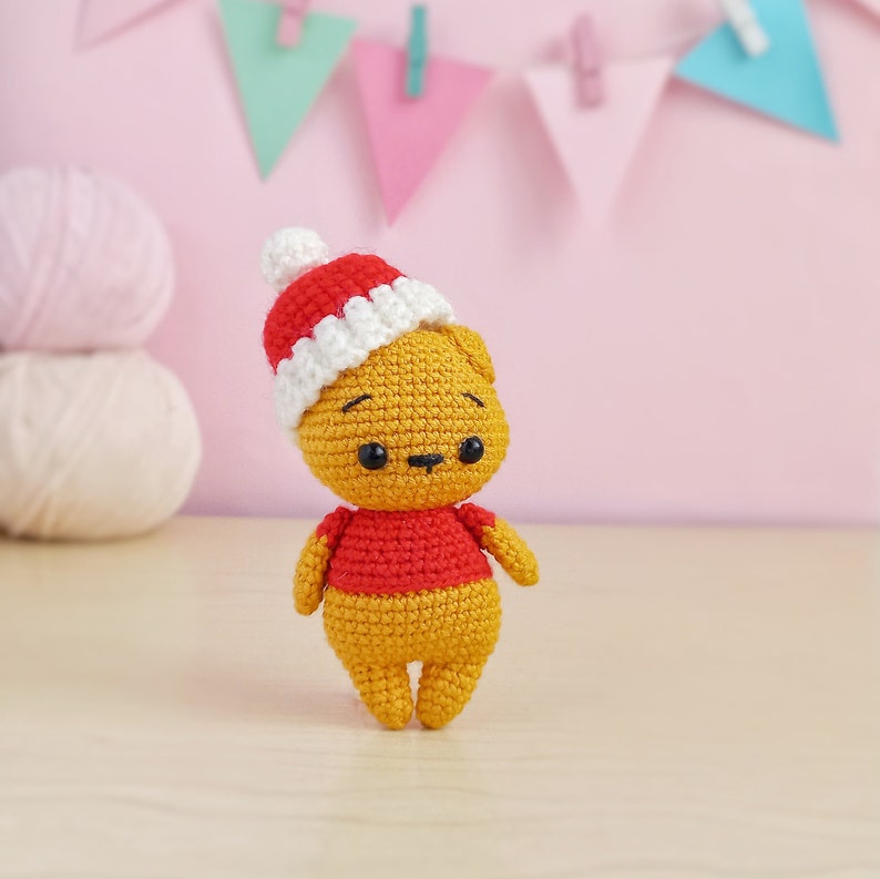 May include: A small crocheted teddy bear wearing a red and white Santa hat. The bear is standing on a light wood surface.