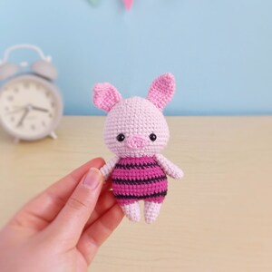 May include: A pink crocheted pig wearing a pink and black striped outfit. The pig has black eyes and a pink snout.