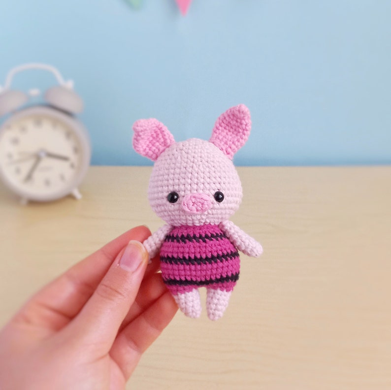 May include: A pink crocheted pig wearing a pink and black striped outfit. The pig has black eyes and a pink snout.