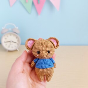 May include: A small crocheted brown elephant wearing a blue shirt. The elephant has pink ears and black eyes. The elephant is being held in a person's hand.