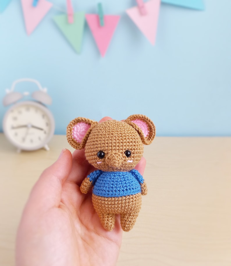 May include: A small crocheted brown elephant wearing a blue shirt. The elephant has pink ears and black eyes. The elephant is being held in a person's hand.