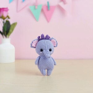 May include: A small, crocheted purple elephant toy with pink ears and black eyes. The elephant is standing on a light wood surface.