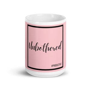 Puede incluir: Taza de cerámica blanca con fondo rosa y borde negro. El texto "Unbothered" está escrito en letra cursiva negra. El hashtag "#PRODUCTIVE" está escrito en negro en la parte inferior de la taza.