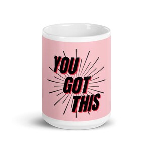 Puede incluir: Una taza de cerámica blanca con fondo rosa y texto negro que dice "YOU GOT THIS" con contorno rojo. El texto está rodeado por un diseño de sol negro.