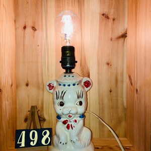 May include: A white ceramic bear lamp with red, blue, and white painted details. The bear is sitting on a base and has a light bulb in its head. The lamp is plugged in and has a cord.