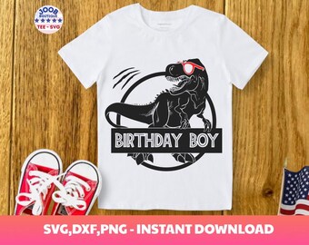 May include: White t-shirt featuring a black dinosaur wearing red sunglasses, with the text "BIRTHDAY BOY" in a black rectangle. The shirt is on a wooden surface, with red sneakers and an American flag in the background.