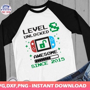 May include: A white and black baseball style shirt with a green and blue video game controller graphic. The text "LEVEL 8 UNLOCKED AWESOME SINCE 2015" is printed on the shirt.