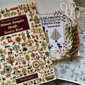 May include: A book titled "The Sampler Company's Ultimate Sampler Motifs Source Book" by Brenda Keyes, featuring a variety of cross-stitch patterns. The book is open to a page with a colourful illustration of a sampler with a house, trees, and animals. There are also several skeins of embroidery floss in green, brown, and orange.