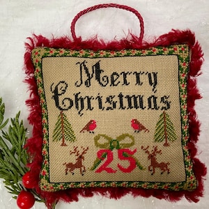 May include: A square Christmas ornament with a burlap-like center featuring the words "Merry Christmas" in black cross-stitch. The ornament has a red fringe border and a red loop for hanging. The design includes trees, birds, reindeer, and the number 25.