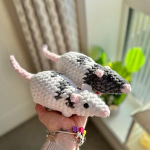 May include: Two handmade crochet plush rats featuring a soft, textured white and dark grey yarn pattern. Each rat has small pink ears, a pink nose, a long pink tail, and black bead eyes. The plush toys are crafted with a chunky, fuzzy yarn material.