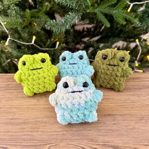 May include: Four handmade crochet frog toys in shades of green and blue. Each frog has black eyes and a stitched mouth. The frogs are displayed on a wooden surface with a blurred background of Christmas tree branches.
