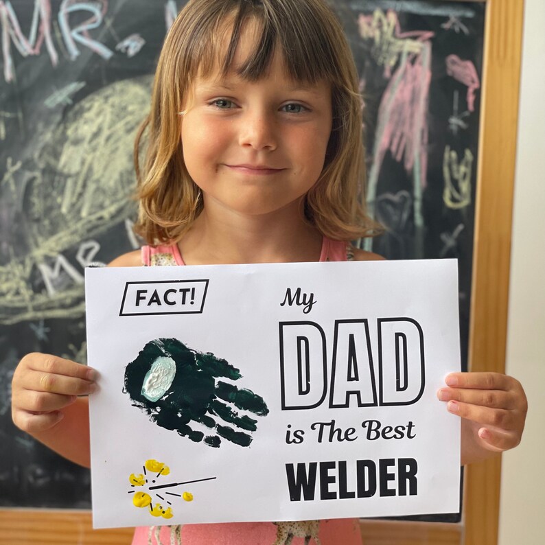 May include: A child holds a white sign with black text that reads "My Dad is The Best Welder." The sign also has a green handprint and yellow sparklers. The child is smiling.