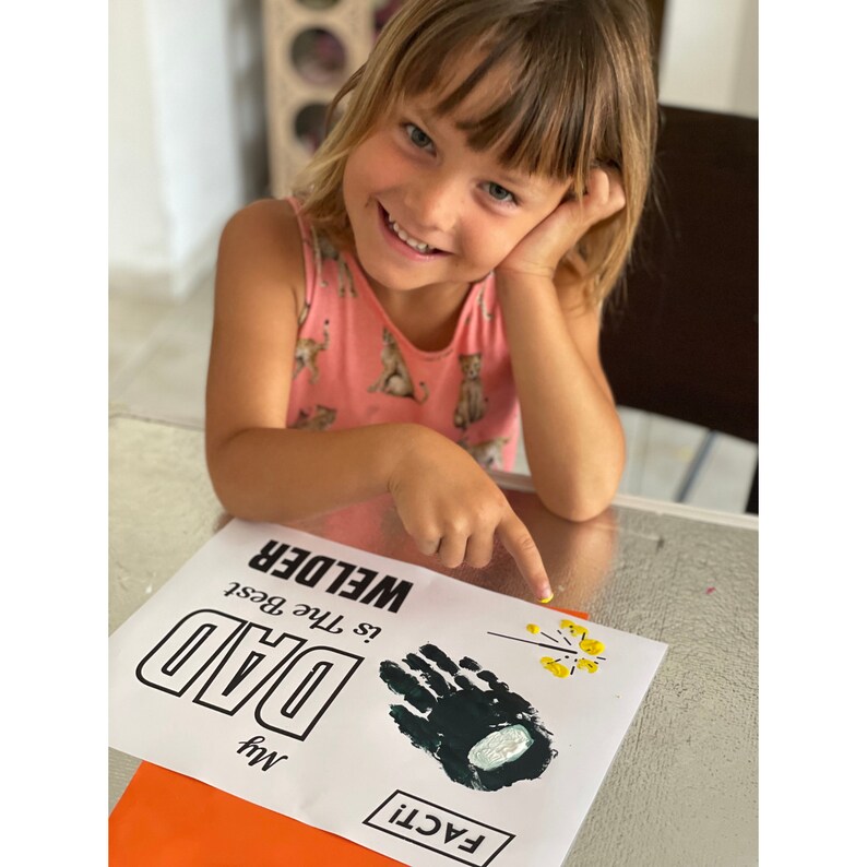 May include: A child holds a white card with black text that reads "My Dad Is the Best Welder". A black handprint is on the card with yellow paint splatters nearby. The card is on a bright orange surface.