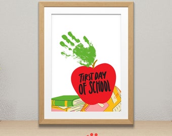 May include: A red apple with the words "First Day of School" written on it. A green handprint is on top of the apple. The apple is sitting on a stack of books.