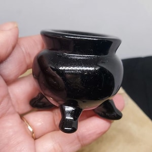 Könnte beinhalten: Ein kleiner, glänzend schwarzer Kessel mit drei kurzen Beinen. Der Miniaturtopf hat einen abgerundeten Körper und eine breite, offene Oberseite. Der Kessel wird in der Hand einer Person gehalten.