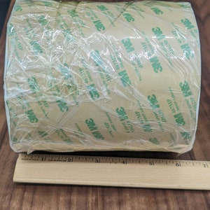May include: A roll of brown 3M tape with green lettering. The tape is wrapped in clear plastic and has a ruler underneath it.