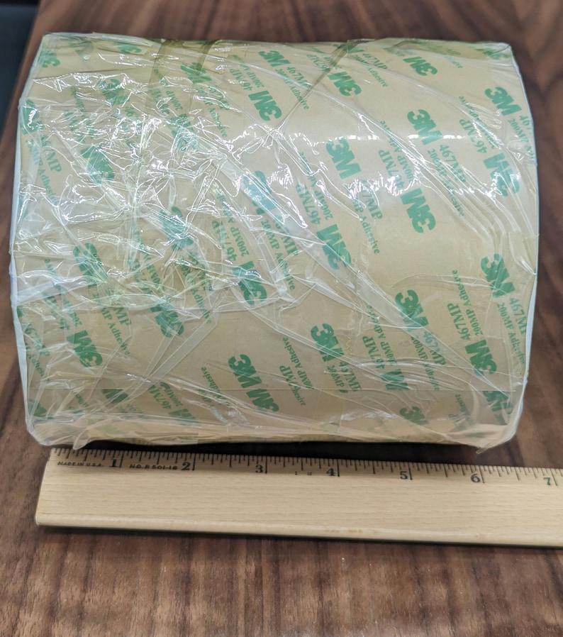 May include: A roll of brown 3M tape with green lettering. The tape is wrapped in clear plastic and has a ruler underneath it.