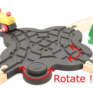 Könnte beinhalten: Ein hölzernes Spielzeug-Zugsset mit einem schwarzen und grauen drehbaren Gleisstück. Das Gleisstück hat einen Knopf, der gedreht werden kann, um die Richtung des Gleises zu ändern. Der Text "Drehen!" ist in Rot auf dem Gleisstück geschrieben.