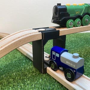 Könnte beinhalten: Ein hölzernes Spielzeug-Zugset mit einer grünen und schwarzen Lokomotive (Nummer 347) und einem blau-grauen Waggon. Die Züge fahren auf Holzschienen und überqueren eine schwarze Brücke auf einer grünen Oberfläche.