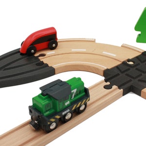 May include: A green toy train with a black roof and a number 7 on the side is on a wooden track. A red toy car is on a curved wooden track with a black and gray section. A green toy tree is in the background.