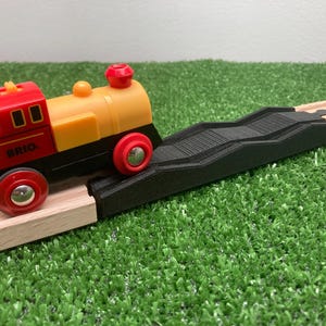 May include: A red, yellow, and black toy train with red wheels is on a wooden track, approaching a black bridge. The train is labeled "BRIO". The scene is set on a green, artificial grass surface.