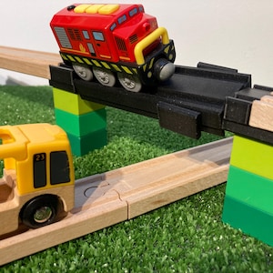 May include: A toy train set featuring a red and yellow train engine on a black bridge, with a yellow bus. The train track is made of wood and the bridge is supported by green and yellow blocks, set on a green surface.