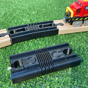 May include: Black plastic train track connectors and a red toy train with yellow accents on a wooden track. The train is positioned on a wooden track, with a black connector piece in front of it. Another connector is in the foreground.