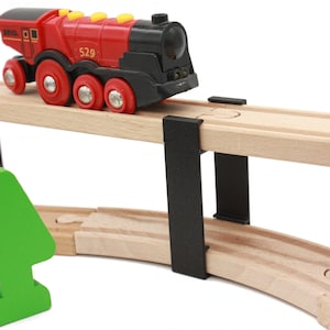 May include: A red and black toy train engine with the number 529 on the side is traveling over a wooden train track bridge. The bridge is supported by two black pillars. A green wooden tree is in the foreground.