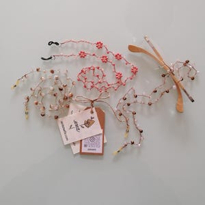 Op de afbeelding: Een bovenaanzicht toont drie glazenkettingen met kralen in roze, bruin en crème. Eén ketting heeft roze bloemvormige kralen. Een bril met houten montuur is versierd met een kralenketting. Labels met de tekst "asraarte" zijn ook zichtbaar.