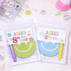 May include: Two clear plastic bags with colorful birthday party favors. Each bag has a smiley face-shaped notepad and a crayon. The bags are labeled "Aria's 8th Birthday" and "Aria's is a Vibe!" with decorative elements.