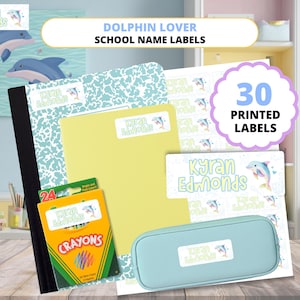 May include: School supplies with dolphin-themed labels. Includes a notebook, folder, pencil case, and a box of crayons. The labels feature a dolphin graphic and the name "Kyran Edmonds". The image also shows a sign that says "30 Printed Labels".