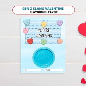 May include: A light blue Valentine's Day card with the text "YOU'RE AMAZING" and candy hearts with Gen Z slang. A round, blue playdough container is at the bottom. Red heart decorations are scattered around the card.