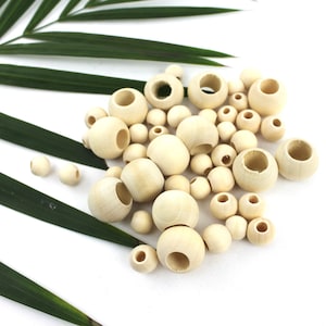 May include: A collection of natural wood beads in various sizes, some with a hole through the centre. The beads are scattered on a white surface with a green palm leaf in the background.